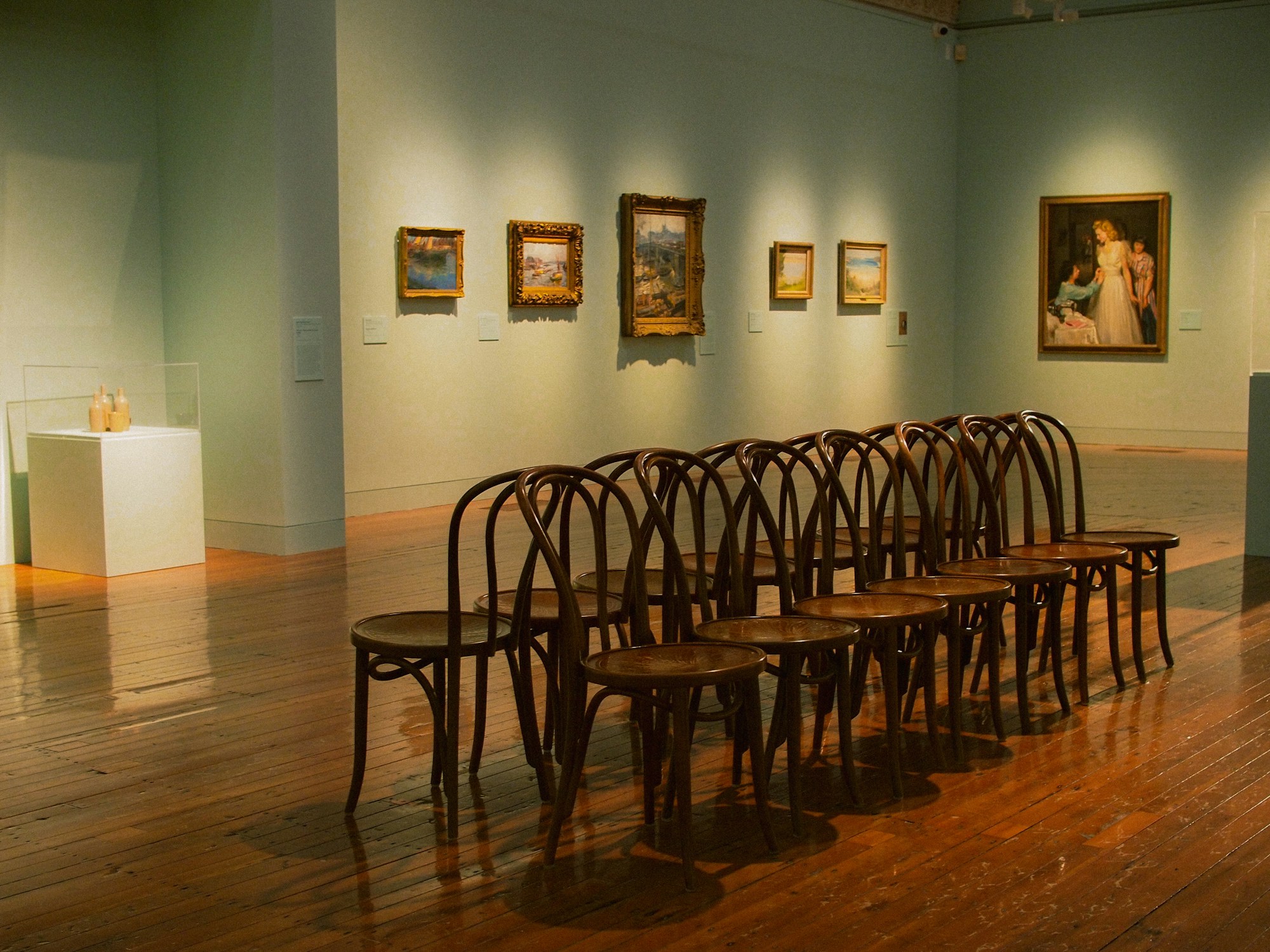 Galeria de arte com pinturas emolduradas, cadeiras de madeira e chão de tábuas iluminado por luz quente.