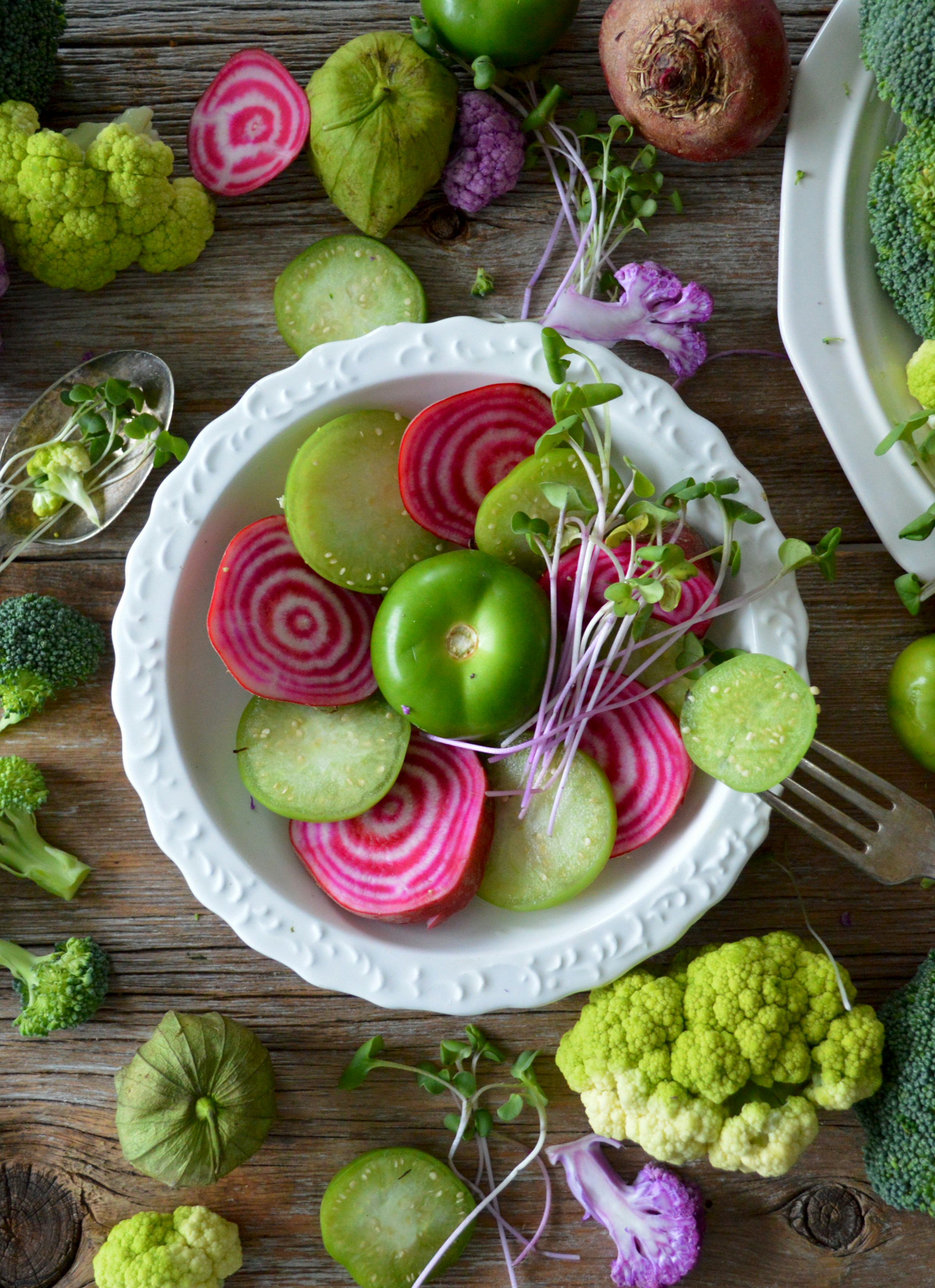 Rodelas de beterraba rosa, hortícolas verdes e rebentos num prato branco, rodeado por brócolos e couve-flor.