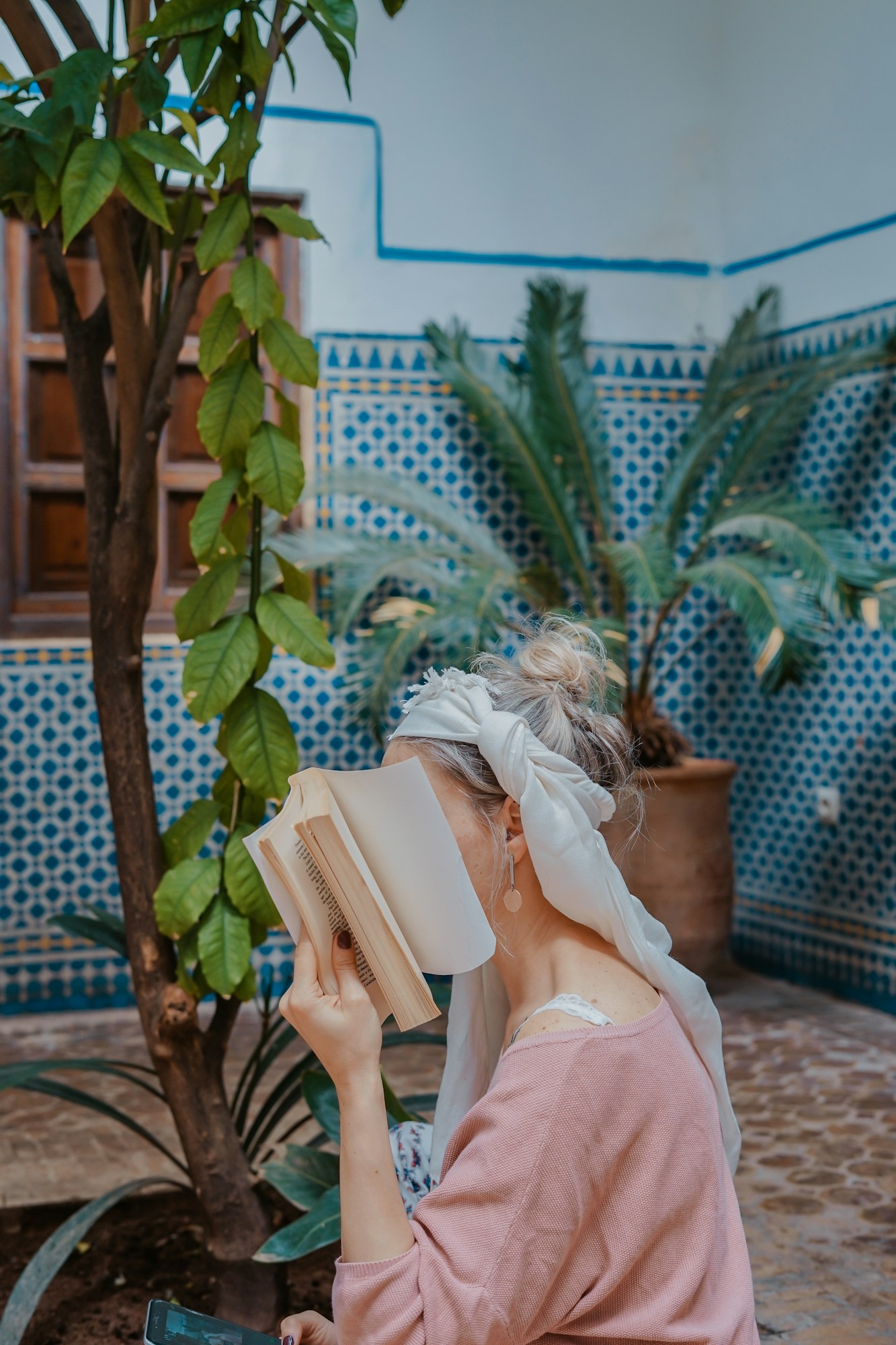 Mulher sentada a ler num pátio com plantas e azulejos azuis.