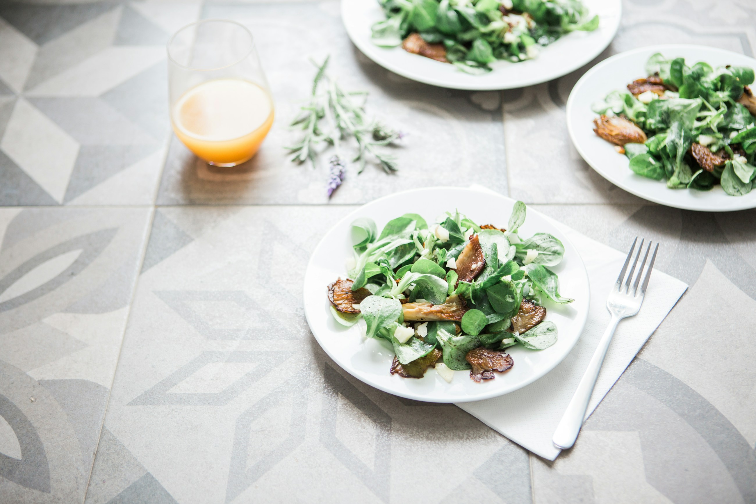 Três pratos de salada de folhas verdes e cogumelos sobre uma mesa clara, com talheres e um copo de sumo.