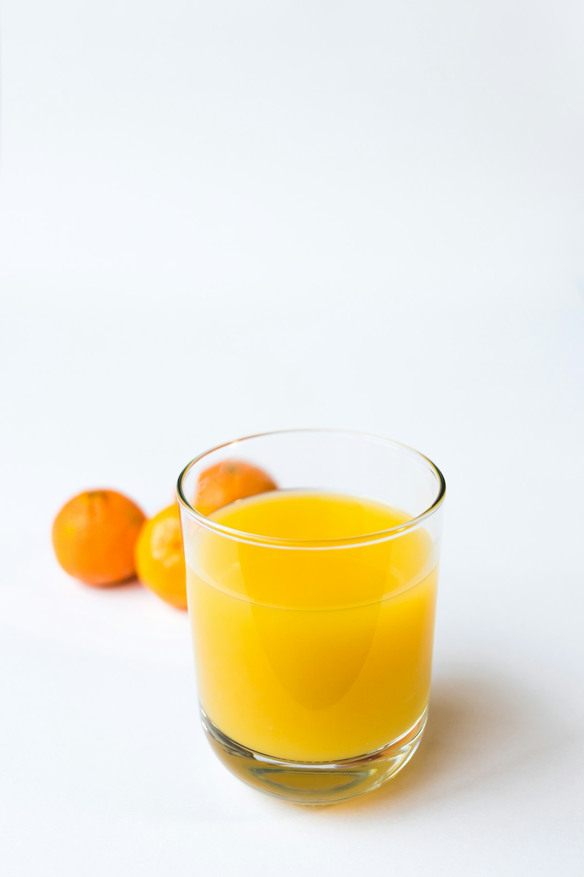 Copo de sumo de laranja, com citrinos ao fundo, sobre uma superfície branca.