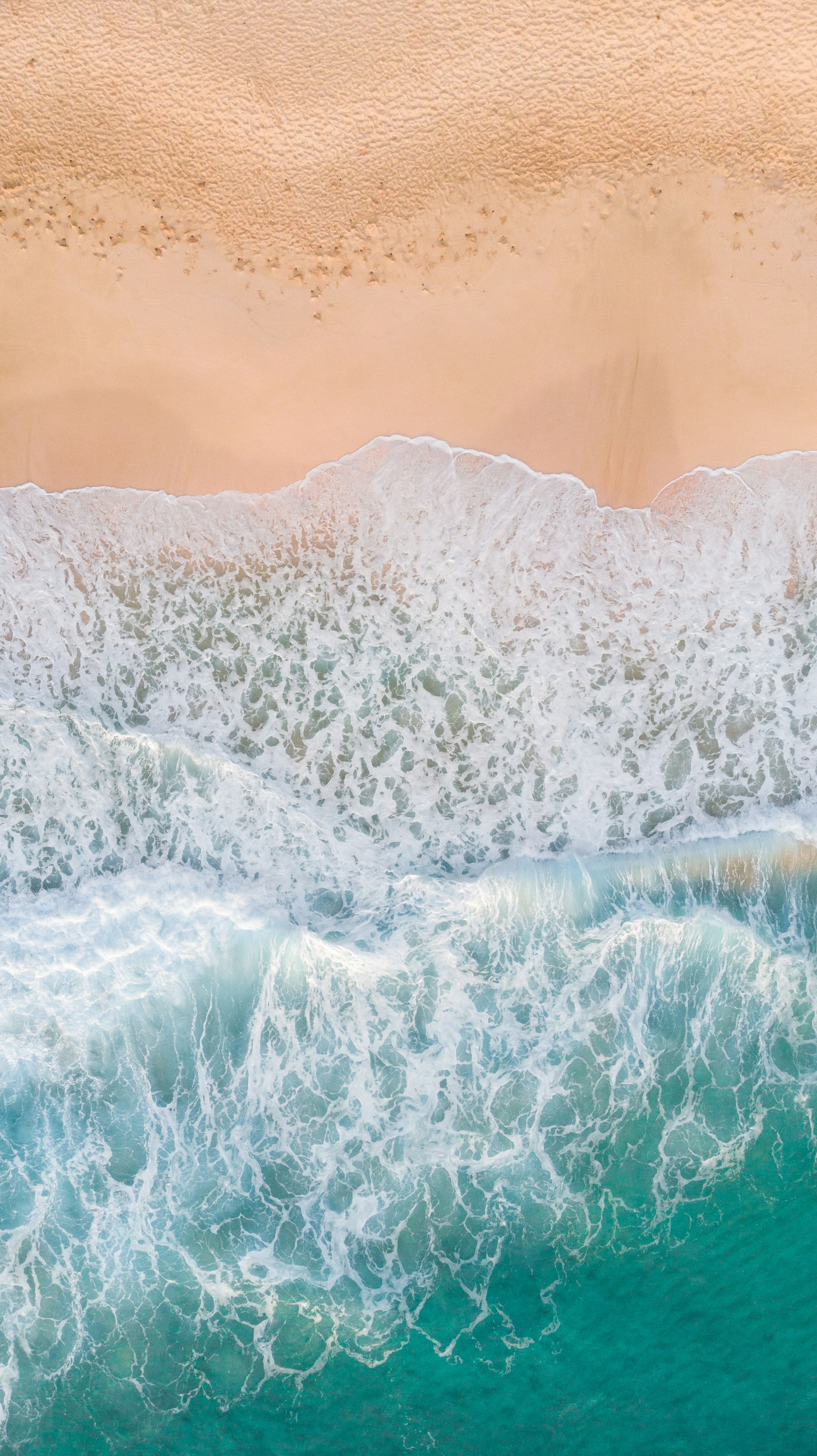 Vista aérea de ondas com espuma branca a chegar a um areal claro, junto a água turquesa.